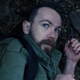 A man in a green Army jacket cowers on the forest floor.
